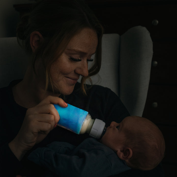 A woman lovingly feeds a baby using a Dr. Brown's Natural Flow® Options+™ Narrow Glass Bottle Silicone Sleeve. They are sitting in a dimly lit room, creating a cozy and intimate atmosphere. The woman gazes warmly at the baby, who is wrapped in a blanket, while the silicone sleeve adds a touch of safety to their serene moment.