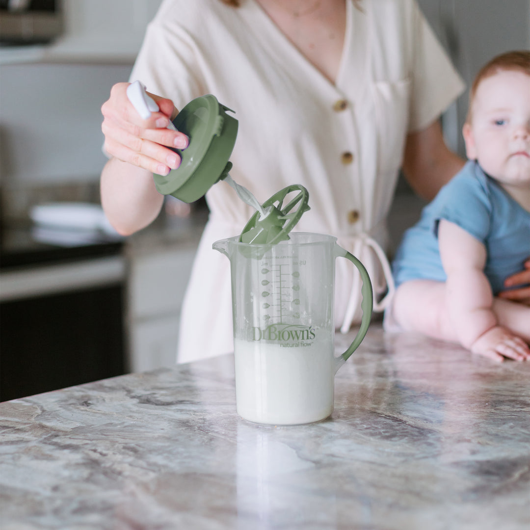 A person expertly using a Dr. Brown's Natural Flow® Formula Mixing Pitcher with a green lid on a marble countertop prepares baby formula while the baby sits nearby. They wear a beige buttoned shirt, ensuring everything is ready for the baby bottles.