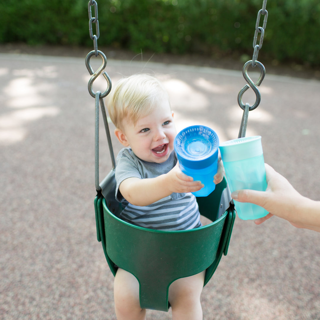 A smiling toddler with blonde hair sits in a swing, wearing a striped shirt and reaching out to take a blue Dr. Brown’s® Milestones™ Cheers360™ Cup, 10 oz/300 mL from an adult's hand. The background features a blurred view of a park with green shrubs.