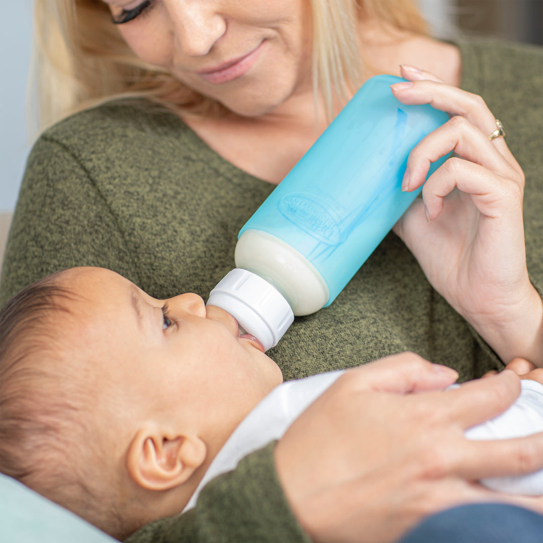 A baby is being bottle-fed by a woman in a green sweater, gently cradling the child while using Dr. Brown's Natural Flow® Options+™ Narrow Glass Bottle with Silicone Sleeves for safety. The scene exudes care and nurturing.