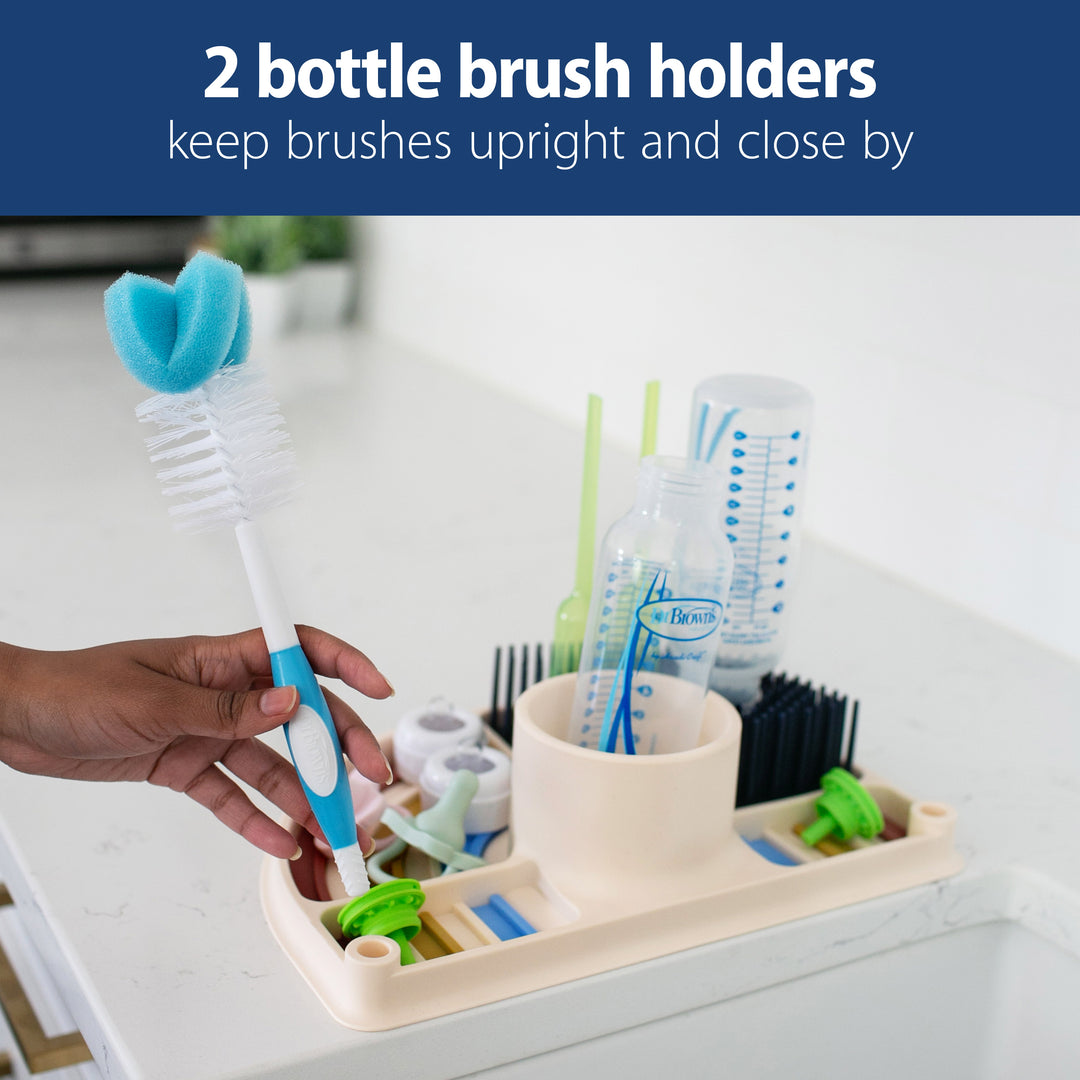 Using the Dr. Brown’s™ 2-in-1 Dry & Prep Silicone Mat, a person places a baby bottle brush into one of the two holders on the drying rack, which is surrounded by baby bottles, cleaning brushes, and small accessories. Above, the text reads, "With 2 bottle brush holders to keep brushes upright and close by," ensuring that every detail of bottle prep is efficient.