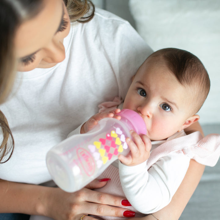 A woman, with bright red nails, is seated comfortably cradling a baby who is drinking from the Dr. Brown's Natural Flow® Anti-Colic Options+™ Wide-Neck Sippy Bottle Starter Kit. Both are wearing white tops in this cozy setting, while the baby gazes up contentedly.