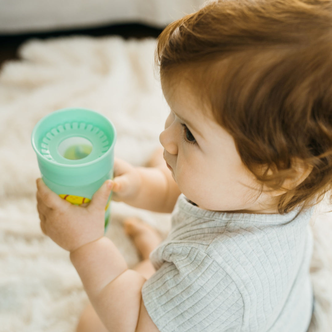 A toddler with light brown hair sits on a soft, cream-colored rug, holding a Dr. Brown's® Milestones™ Cheers360™ Cup 10 oz/300 mL in both hands. The child is wearing a short-sleeved, light gray shirt and gazes to the side, enjoying the spoutless design that allows for easy sipping from any angle.