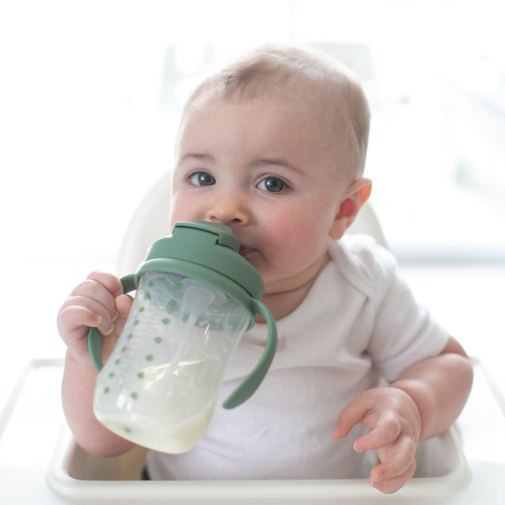 A baby in a high chair sips from Dr. Brown's™ Milestones™ Baby’s First Straw Cup, which features a spill-proof design with green handles and lid. The little one, dressed in a white shirt, gazes toward the camera with curiosity. Soft natural light bathes the scene as the weighted straw ensures every sip is delightful for the baby.