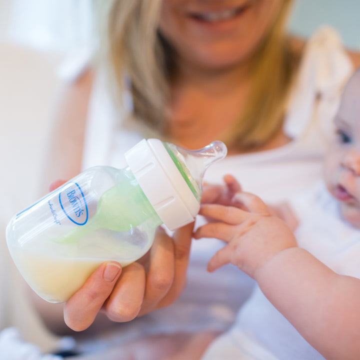 A woman holds a baby in her arms as the little one reaches for a Dr. Brown’s Natural Flow® Wide-Neck Baby Bottle filled with milk. This tender moment, enhanced by the bottle's silicone nipple from Dr. Brown's, beautifully captures the nurturing and caring bond between them.