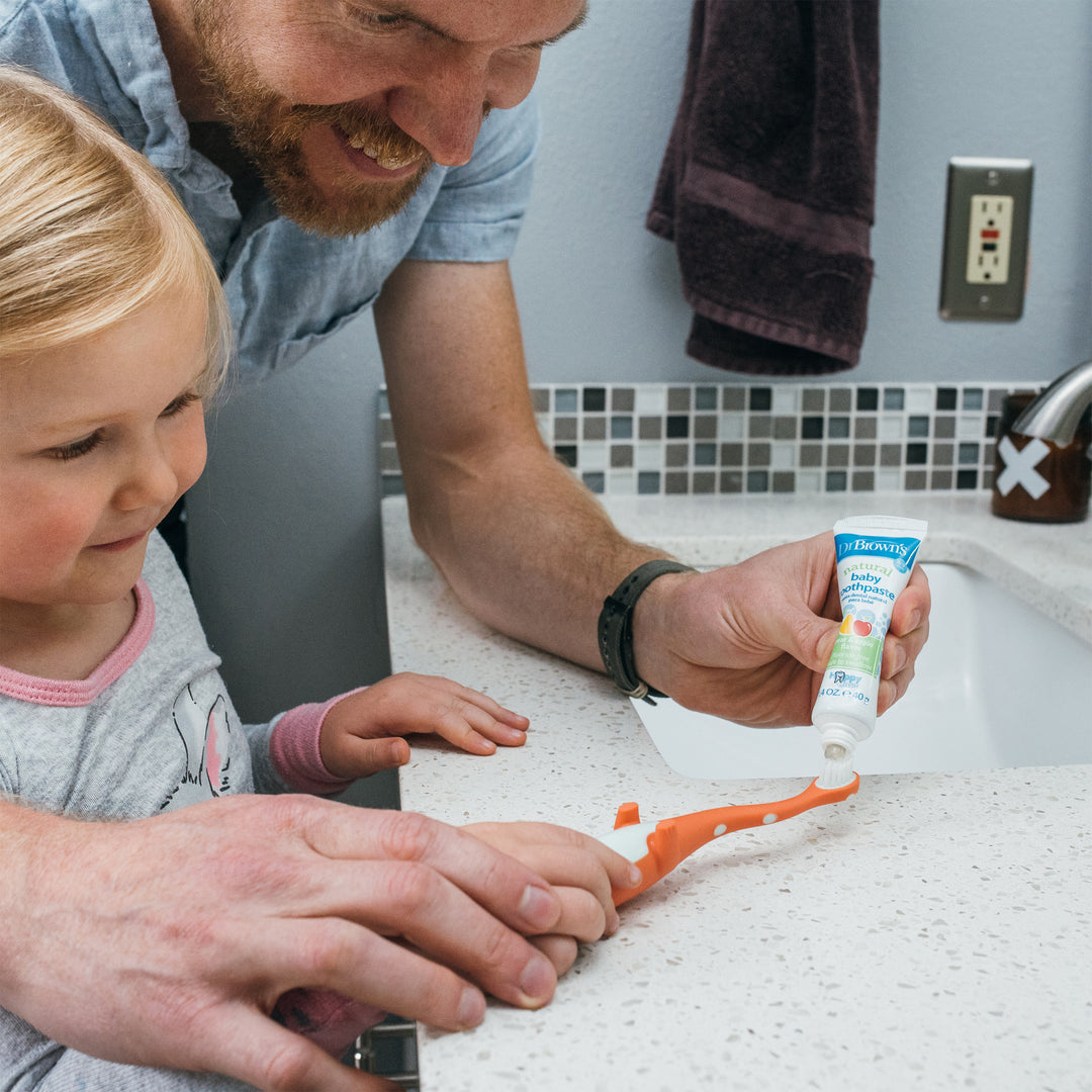 A smiling man helps a young child apply Dr. Brown’s™ Fluoride-Free Baby Toothpaste, Apple Pear flavor, to their toothbrush. Beside a modern sink with a light-colored countertop, they embrace dental hygiene habits together, striving for clean teeth.