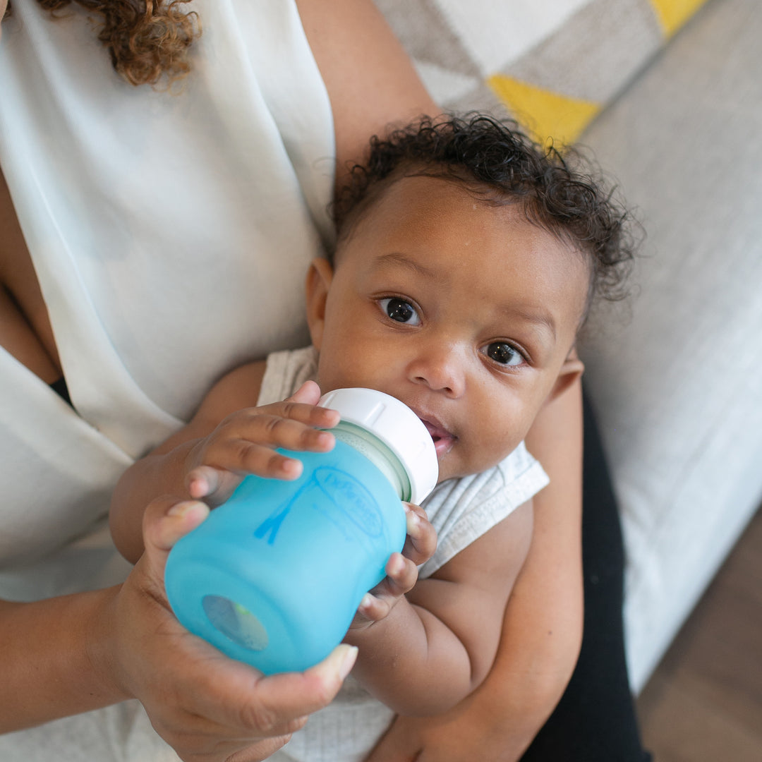 A baby with curly hair holds a blue glass bottle featuring Dr. Brown's Natural Flow® Options+™ Wide-Neck Glass Bottle Silicone Sleeves while sitting on a woman's lap. The woman's torso and arm are visible as she supports the baby, and they appear to be indoors on a couch with a colorful blanket.