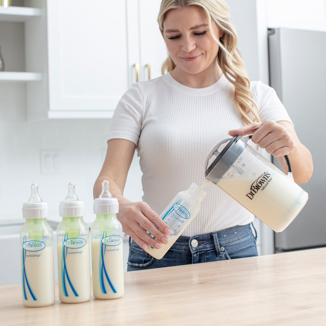 A woman in a white shirt and jeans effortlessly pours milk into a baby bottle using Dr. Brown's Natural Flow® Formula Mixing Pitcher, aiming to reduce gas for her baby. On the kitchen countertop, three other filled baby bottles are neatly arranged against the sleek white cabinets.