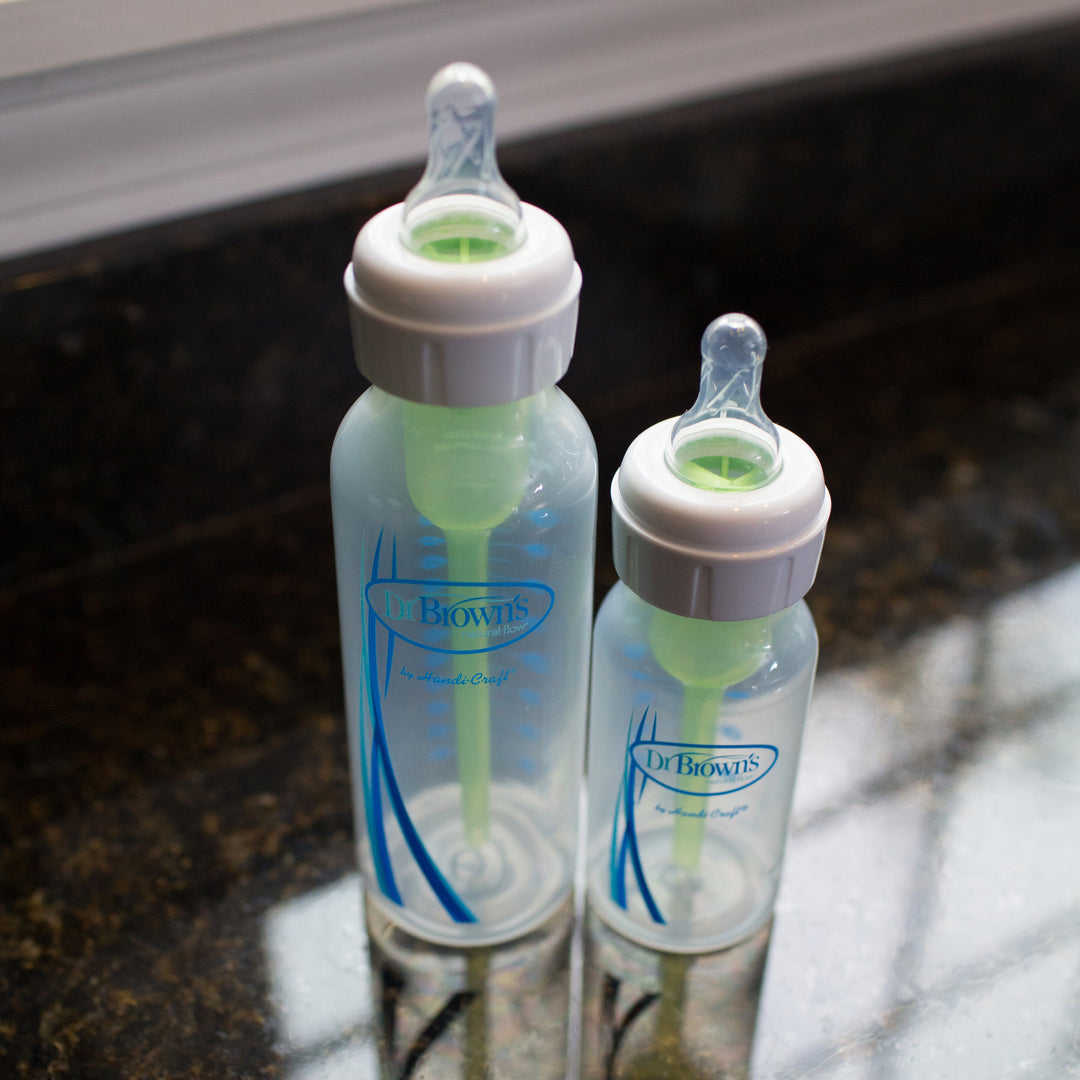 Two Dr. Brown's baby bottles with green internal vents sit on a dark countertop. One narrow bottle is larger than the other, both featuring clear bodies and white caps. Light reflects off the surface, casting a soft glow around the bottles fitted with Dr. Brown’s™ Medical – Narrow Bottle Nipples.